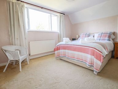 A bedroom with a bed with striped bedding two plaid pillows a white wicker chair near a window and cream carpet at Trevow in Camborne