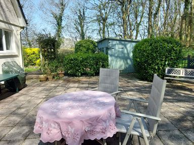 An outdoor patio with a round table covered by a pink tablecloth and two chairs near a light blue garden shed and bushes at Trevow in Camborne