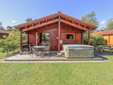 A wooden cabin with a covered porch featuring a table with chairs and a hot tub in a grassy yard at Jemima Lodge in Tattershall