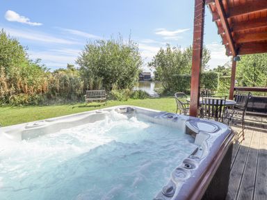 An outdoor hot tub with bubbling water next to wooden deck with table and chairs and greenery at Jemima Lodge in Tattershall