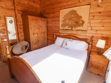 A bedroom with a wooden bed white linens a wooden wardrobe a bedside table with a lamp a wicker chair and a wall hanging of a tree at Jemima Lodge in Tattershall