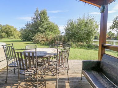An outdoor patio with a round metal table and six chairs on a wooden deck overlooking a grassy area with bushes and trees at Jemima Lodge in Tattershall