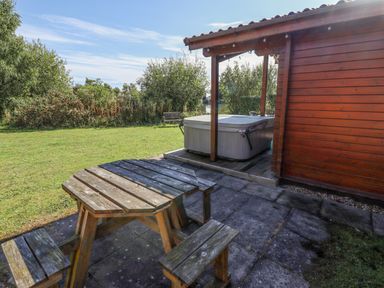 A wooden picnic table and benches on a patio next to a hot tub and wooden cabin with grass and trees in the background at Jemima Lodge in Tattershall