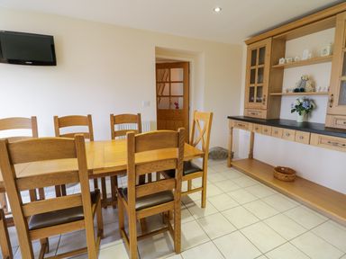 A dining room with a wooden table and chairs a wooden cabinet with shelves and a television mounted on the wall at Tyn Y Buarth in Dyffryn Ardudwy