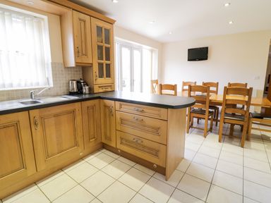 A kitchen with wooden cabinets a black countertop a dining table with six wooden chairs and a wall-mounted television at Tyn Y Buarth in Dyffryn Ardudwy