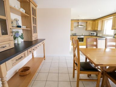 A kitchen with wooden cabinets a dining table with chairs and a countertop with shelves and decorations at Tyn Y Buarth in Dyffryn Ardudwy