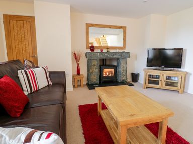 A living room with a brown leather sofa red and striped cushions a wooden coffee table a stone fireplace with a fire and a flat screen TV on a wooden stand at Tyn Y Buarth in Dyffryn Ardudwy