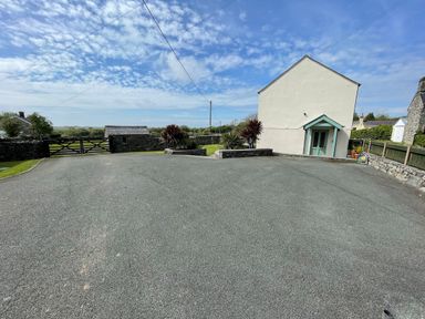 A paved driveway leading to a small white house with a green door and a stone wall with plants on either side at Tyn Y Buarth in Dyffryn Ardudwy