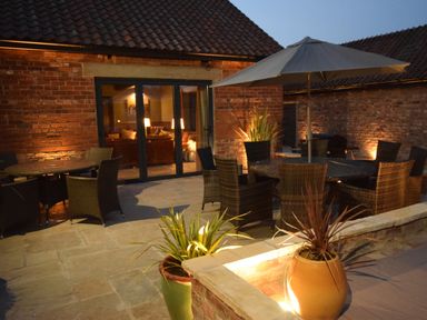 An outdoor patio with wicker chairs tables an umbrella large potted plants and a brick house with glass doors at Sykes Lodge in Malton
