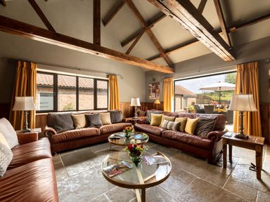 A living room with brown leather sofas, cushions, glass coffee tables with flowers, lamps, wooden beams, and yellow curtains at Sykes Lodge in Malton