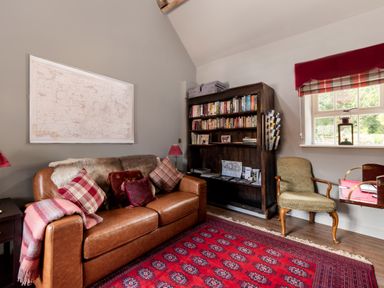 A room with a brown leather sofa with cushions and a blanket a wooden bookshelf a green upholstered chair and a window with a red valance at Sykes Lodge in Malton