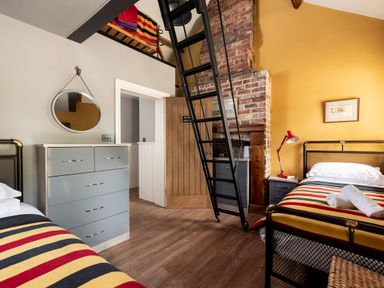 A bedroom with two single beds striped blankets a gray chest of drawers a round mirror a black ladder leading to a loft and a yellow painted wall at Sykes Lodge in Malton