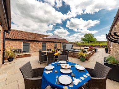 An outdoor patio area with dining tables and chairs under an umbrella beside a hot tub next to brick buildings