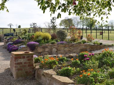 A flower garden with stone walls and gravel pathways overlooking a fenced green field at Sykes Lodge in Malton