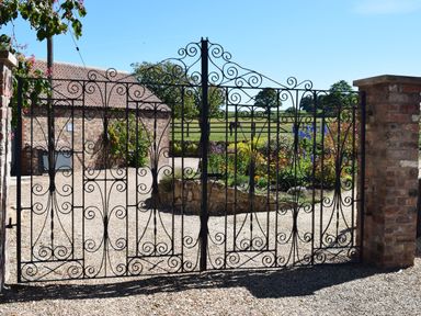 An iron gate with decorative patterns in front of a gravel driveway with brick pillars and a garden behind at Sykes Lodge in Malton