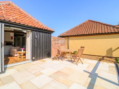 An outdoor patio with a wooden table and four chairs next to a black wooden building with open doors showing a living room at Sparrow Barn in Reepham