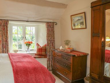 A bedroom with a bed covered in red blanket near a window with floral curtains and a wooden dresser with a painting above at Rookwood in Drewsteignton