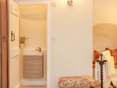 A small sink with a wooden cabinet in a beige room with a floral bench and a chair with blankets and red checkered cushions at Rookwood in Drewsteignton