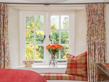 A window with floral curtains a vase of flowers and a plaid cushion on the window seat at Rookwood in Drewsteignton