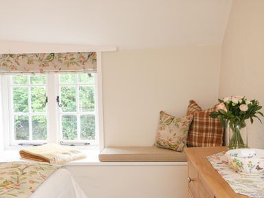 A window with floral curtains and a window seat with cushions and a folded blanket next to a wooden dresser with flowers in a vase at Rookwood in Drewsteignton