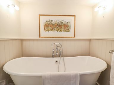 A bathroom with a white freestanding bathtub a towel hanging on the tub a framed picture above the tub and wall lights at Rookwood in Drewsteignton