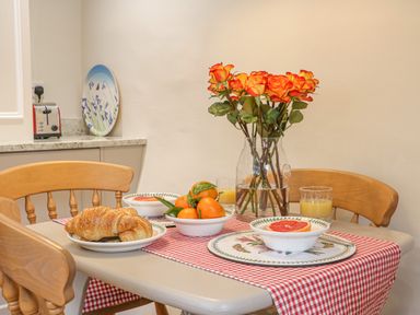 A kitchen table with a red and white checkered table runner orange flowers in a vase croissants bowls of fruit and glasses of juice at Rookwood in Drewsteignton