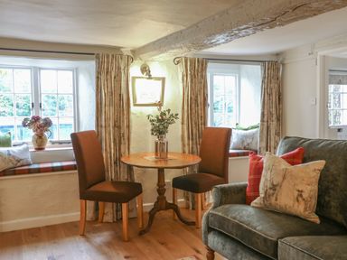 A room with a small round wooden table and two brown chairs next to two windows with curtains and a green sofa with pillows at Rookwood in Drewsteignton