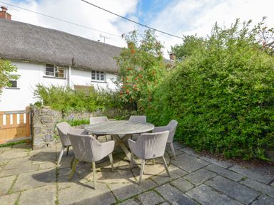 An outdoor patio area with a round wooden table and six wicker chairs surrounded by bushes at Rookwood in Drewsteignton