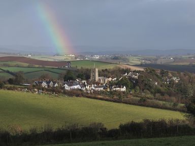 A village with houses and a church tower surrounded by fields and a rainbow in the sky at Rookwood in Drewsteignton