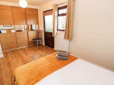 A bedroom with a bed covered in a white sheet and orange blanket a wooden wardrobe with mirrored panels a wooden chair and a door with a window at Daffodil Lodge in Saltburn-by-the-Sea