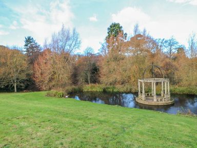 A grassy area with a small pond and a stone gazebo surrounded by trees at Orchid Lodge in Saltburn-by-the-Sea