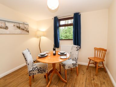 A dining area with a round wooden table set for two with floral-covered chairs a wooden chair in the corner a tall lamp and a window with blue curtains at Foxglove Lodge in Saltburn-By-The-Sea