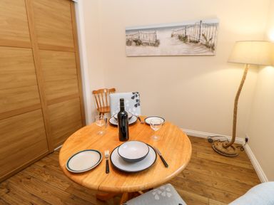 A dining area with a round wooden table set for three with plates glasses and a bottle of wine a floor lamp wooden floor and a beach painting on the wall at Foxglove Lodge in Saltburn-By-The-Sea