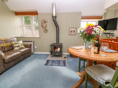 A living room with a brown leather sofa a wood burning stove a round wooden table with flowers and wine glasses at Higher Lampra in Mullion