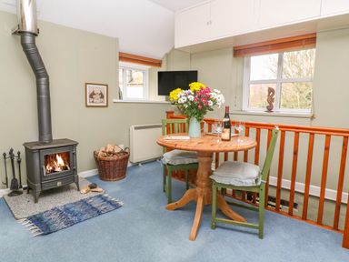 A dining area with a round wooden table with flowers and wine glasses near a wood stove with fire at Higher Lampra in Mullion