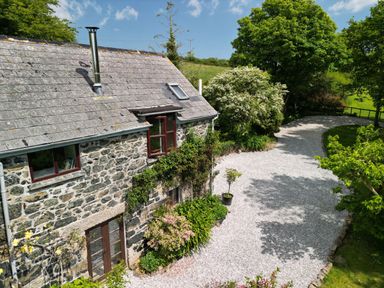 A stone house with a slate roof next to a gravel driveway and surrounded by trees and bushes at Higher Lampra in Mullion