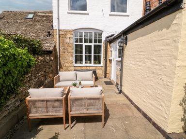 An outdoor patio with wooden chairs, a sofa, and a small table with a plant and a bottle in a courtyard at The Cottage at Broadway in Broadway