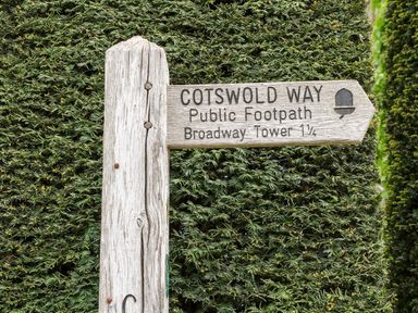 A wooden signpost on a hedge background pointing to Cotswold Way Public Footpath Broadway Tower at The Cottage at Broadway