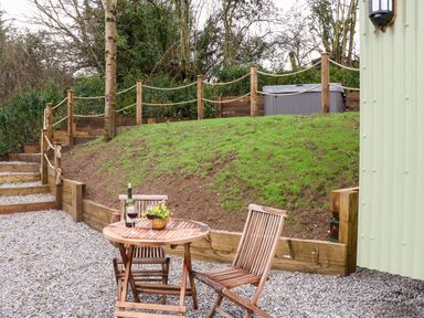 An outdoor patio area with a wooden table and two chairs a bottle of wine and two glasses on the table grass hill with rope railing and steps leading up to a hot tub at Little Silver Leaf in High Bickington