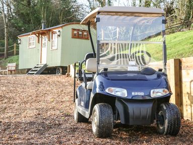 A golf cart parked on a wood chip path next to a green shepherd hut with windows and stairs in a wooded area at Little Silver Leaf in High Bickington