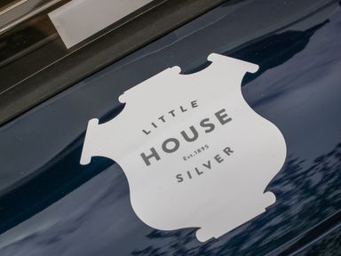 A close up of a shield shaped sign with the text Little House Silver Est 1895 on a dark reflective surface at Little Silver Owl in High Bickington