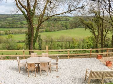 An outdoor seating area with wooden table and chairs on a gravel surface overlooking a countryside landscape with trees and fields at Little Silver Owl in High Bickington