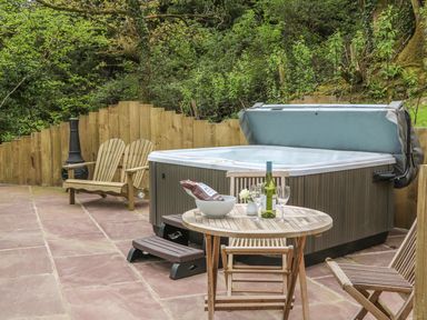 An outdoor patio with a hot tub wooden chairs a round table with wine and glasses and a wooden fence with trees behind at Little Silver Owl in High Bickington