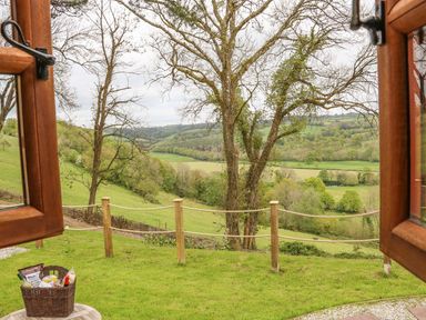 View through open wooden windows of a grassy area with a rope fence and trees overlooking hills and fields at Little Silver Owl in High Bickington