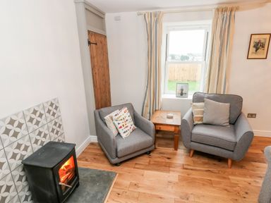 A living room with two gray armchairs a wooden side table a window with striped curtains and a wood burning stove at Coeden Bach in Burry Port