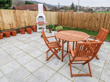 A patio area with wooden table and chairs terracotta pots and a white outdoor barbecue at Coeden Bach in Burry Port
