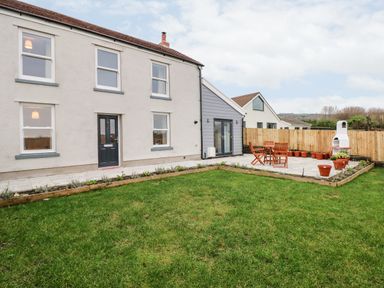 A backyard with green grass wooden patio furniture potted plants a white barbecue and a two-story house at Coeden Bach in Burry Port