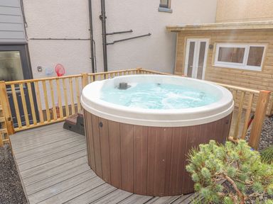 An outdoor hot tub on a wooden deck with a wooden railing and a building with white doors and a window in the background at Coeden Bach in Burry Port