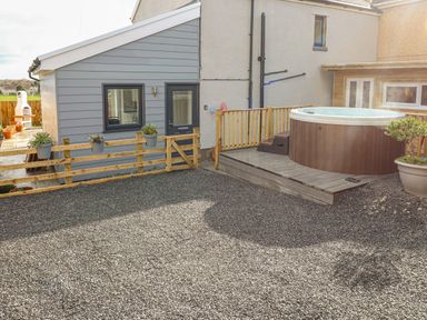 An outdoor area with a hot tub on a wooden deck next to a small house with grey siding and a gravel ground at Coeden Bach in Burry Port
