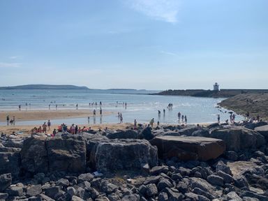 A beach with people on the sand and in the water with a lighthouse on a rocky jetty at Coeden Bach in Burry Port